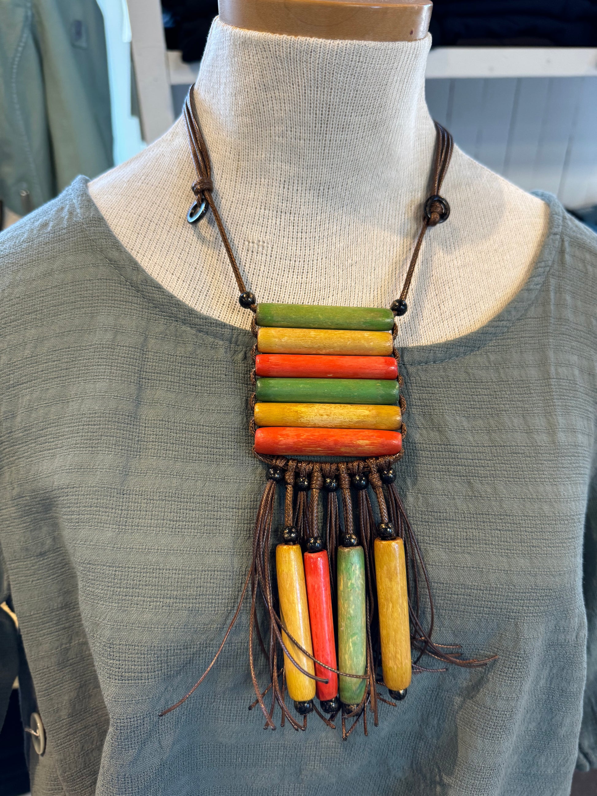 A mannequin dressed in a light gray top showcases the Tropics Adjustable Necklace by Suzie Blue. This distinctive piece features an array of horizontal and vertical beads in vibrant green, yellow, and red hues, perfect for adding a touch of tropical flair. Several beads dangle elegantly at the bottom, all beautifully strung on dark brown cords.