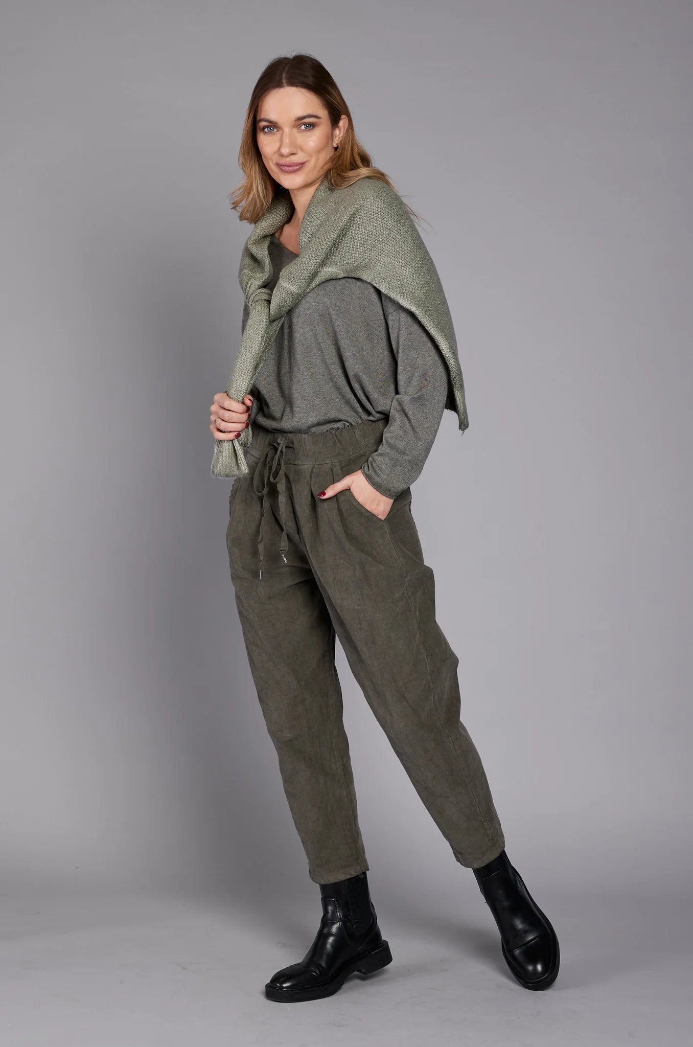 A person wearing a gray long-sleeve top, the stylish EL27901 pants by ELISSIA in ankle-high olive green with a drawstring, and black ankle boots stands with their hands in their pockets against a plain grey background. A scarf is draped over their shoulder. Only the lower half of the person is visible.