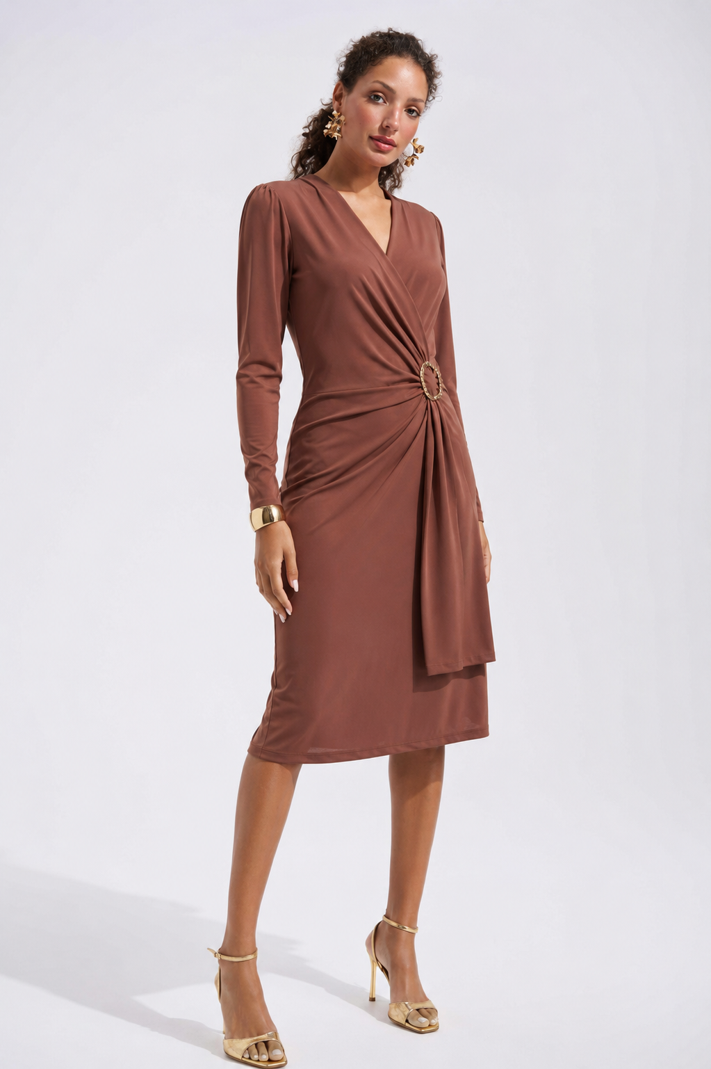 A woman wearing the Joseph Ribkoff Wrap Front Belted Dress Style 233119—a sophisticated long sleeve brown midi dress featuring a gathered waist with a gold buckle—paired with gold earrings, a bracelet, and heels, stands against a light background.