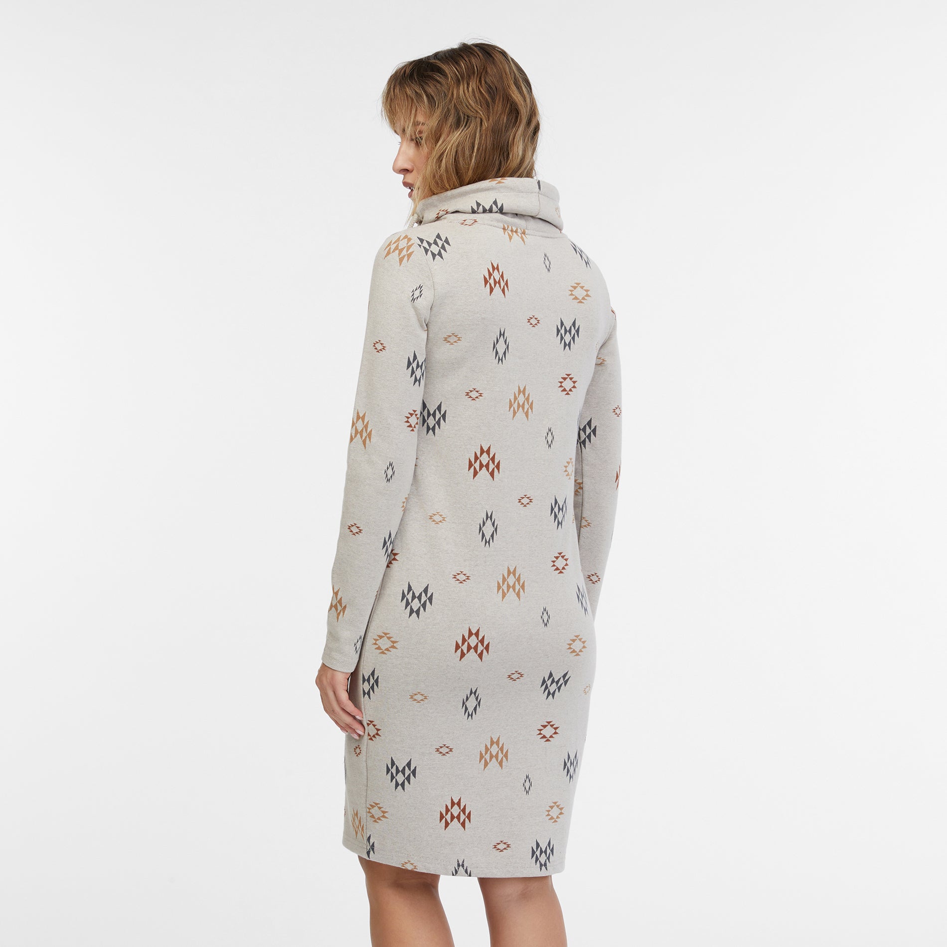 A person with shoulder-length hair is wearing the Sweater Dress Chloenette Ecru by RAGWEAR, a light-gray long-sleeve dress adorned with a geometric diamond pattern in various colors. The dress features a high, loose neck with a drawstring. The background is plain white.