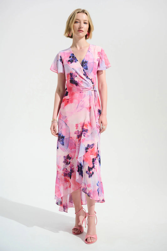 A woman with short blonde hair wears the Joseph Ribkoff Signature Chiffon Print Wrap Dress With Ruffle Detail 261739—pink floral, short sleeves, flowing skirt—paired with pink strappy heels against a plain white background.