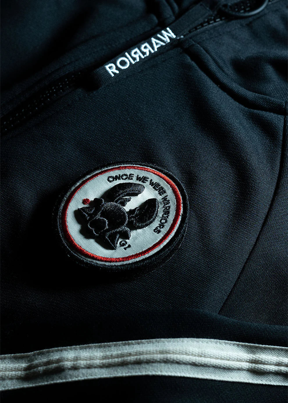 A person with short, curly hair stands in a dark, minimalist studio wearing the Once We Were Warriors Haku Track Jacket - Black and matching pants, which are both adorned with white stripes down the sleeves and legs. The sportswear set features a circular emblem on the chest and thigh. They are also wearing black sneakers and looking downward.