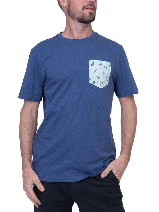 A person stands wearing the Men’s Cotton T-Shirt - BLUE from Silver Jeans Co. The blue, short-sleeved T-shirt features a light blue front pocket with a floral pattern. They gaze to the side with one hand in their black pants pocket, embodying the epitome of casual style against a white background.