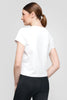 A woman with brown hair in a ponytail is wearing the UCHUU white cotton spandex t-shirt (Model: TOP CS25-303 UCHUU) and black pants, facing slightly to the side against a plain gray background.