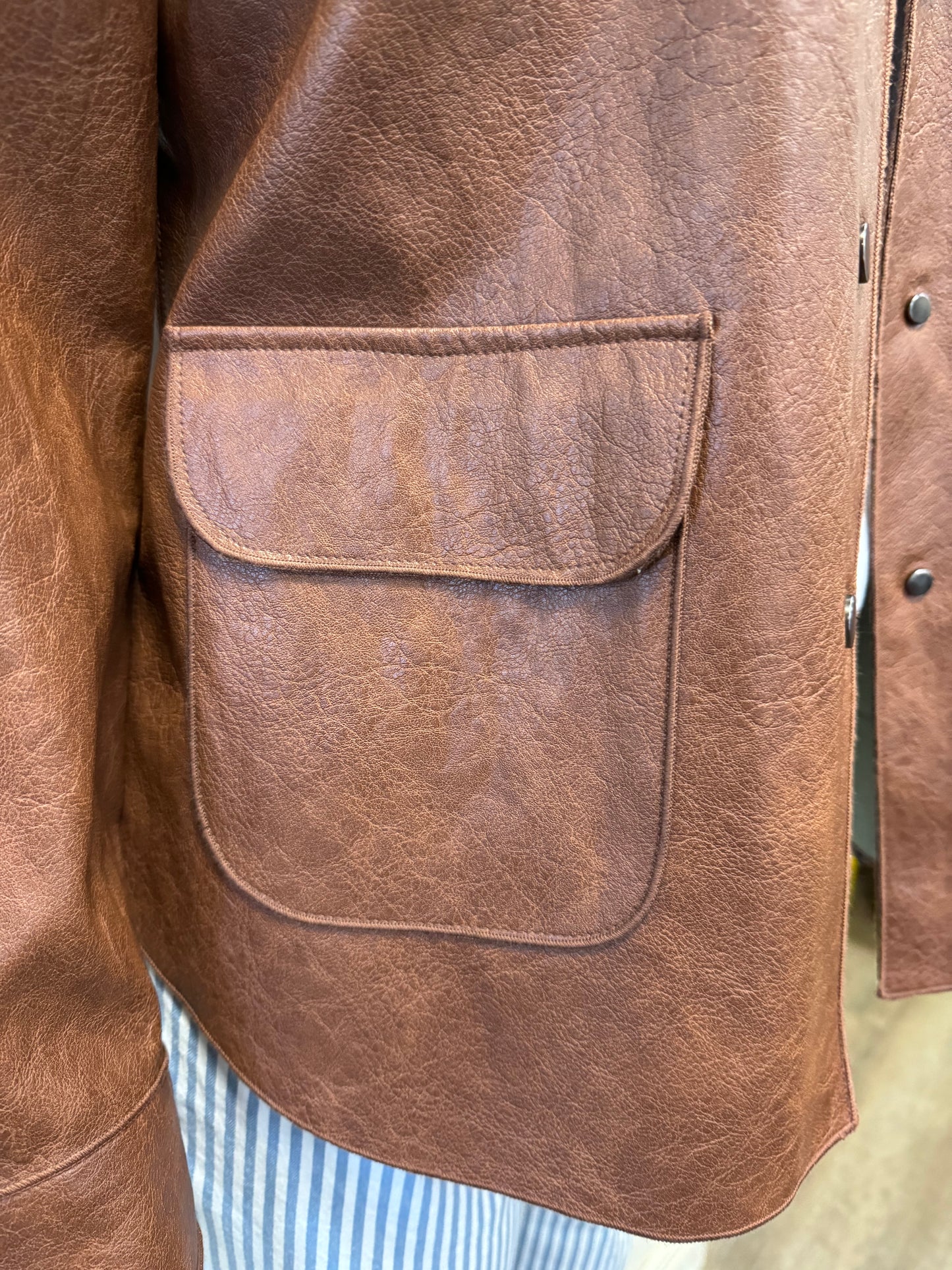 A mannequin wears a stylish and durable Brown Mock Leather Jacket by Tribal over a white T-shirt. The jacket features a collar, buttons down the front, and two large pockets. The mannequin also sports blue and white striped pants. The background is plain and off-white, highlighting the modern design of the outfit.