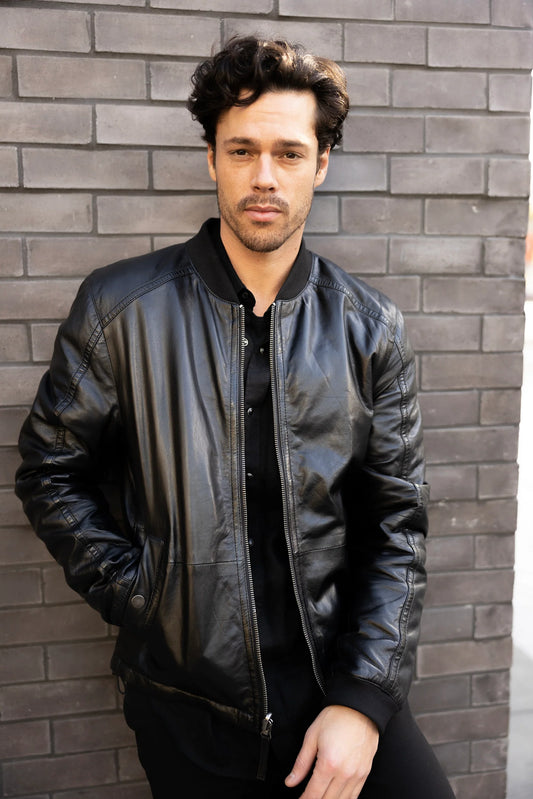 A person with dark, wavy hair and facial hair stands against a brick wall. They are wearing a Cranch CF Leather Jacket, Black - Reversible by MAURITIUS LEATHER, made from lambskin leather over a black shirt, with one hand in the jacket pocket and maintaining a neutral expression.