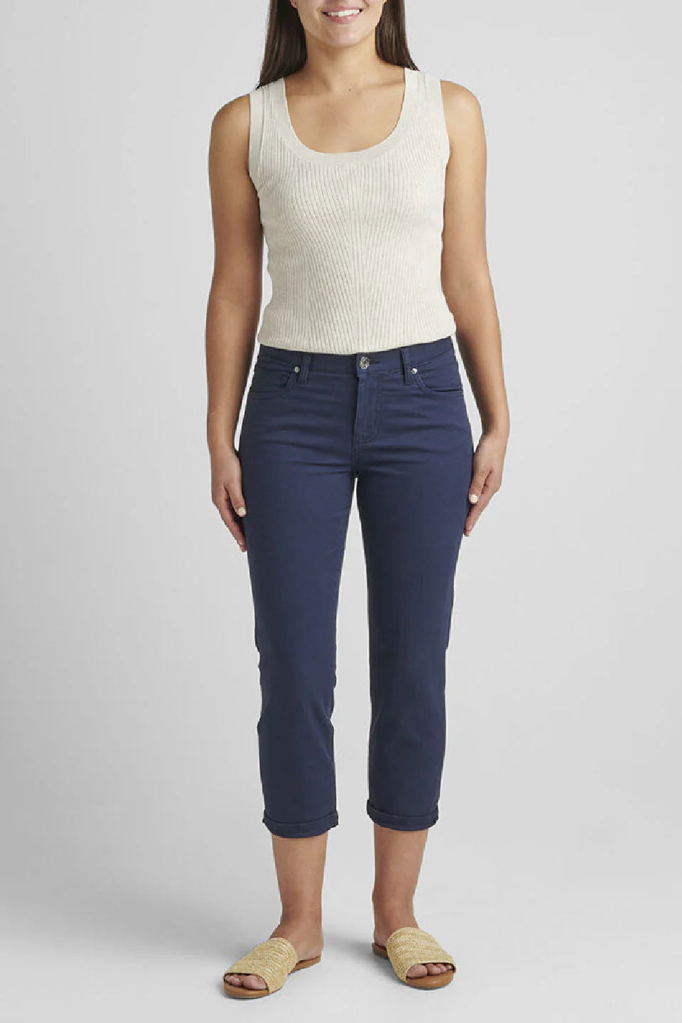 A woman stands against a plain background wearing a sleeveless, white ribbed top, the Cecilia Mid Rise Capri Navy from JAG made from super stretch twill fabric, and beige woven sandals. She has long hair and is facing forward with her arms relaxed by her sides.