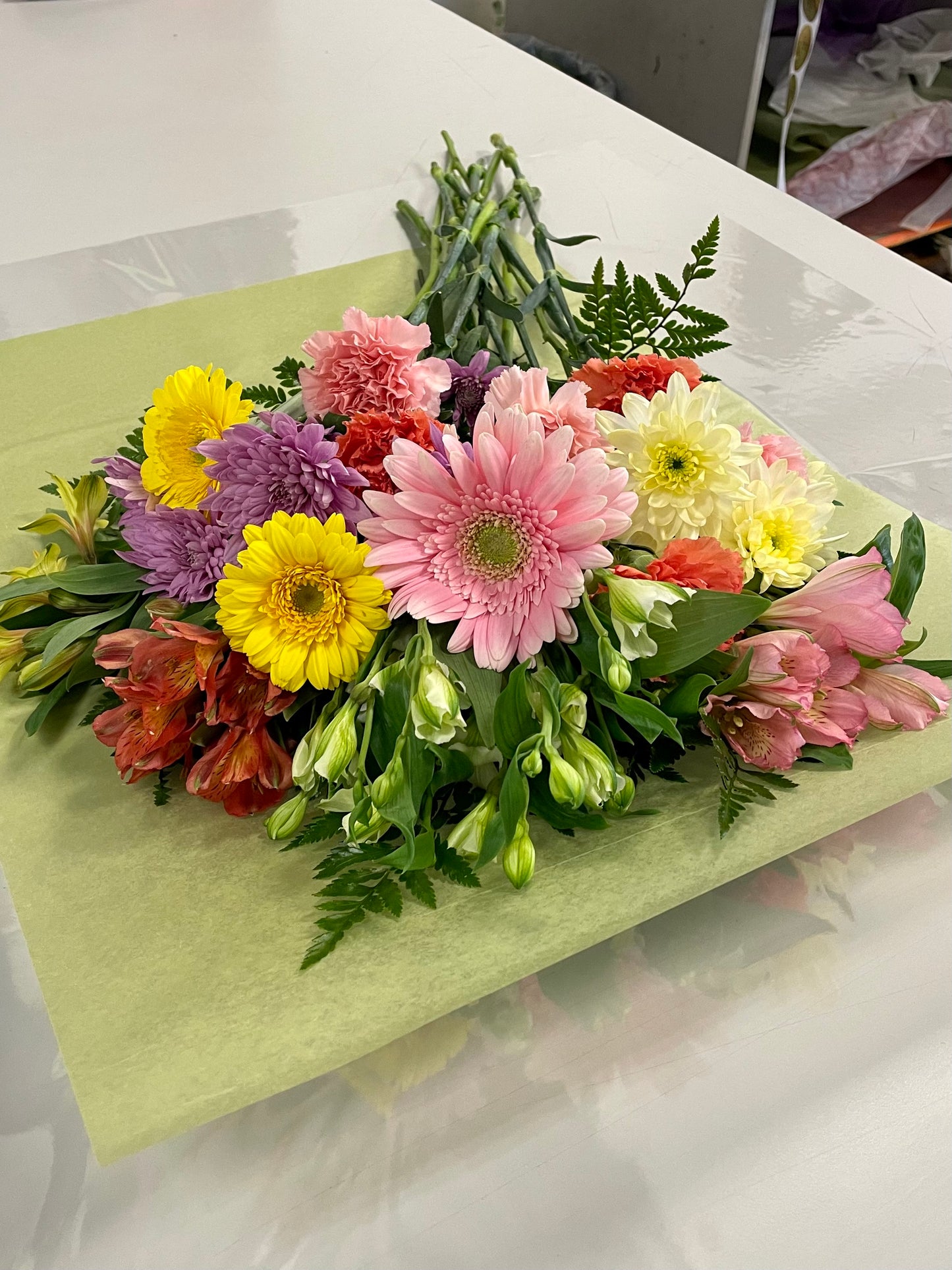 A stunning bouquet called Designer’s Choice Freshcuts from Posies Flowers & Fashion is resting on a white surface. The arrangement features vibrant orange and pink roses, cheerful yellow chrysanthemums, elegant white flowers, and assorted green foliage. It is partially wrapped in brown paper and elegantly tied with a burgundy ribbon.