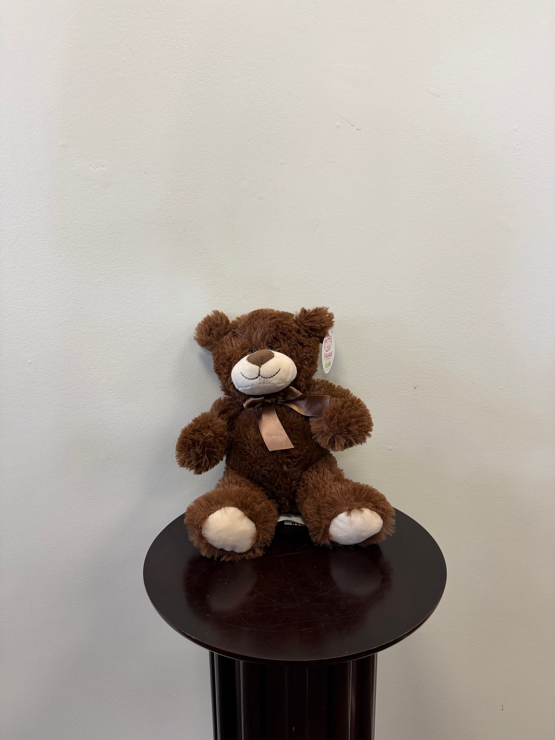 A Classic Teddy Bear by Fiesta, featuring a ribbon around its neck, sits on a dark round pedestal against a plain light-colored wall.