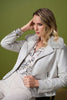 A woman with wavy blonde hair sits against a green backdrop, wearing the Joseph Ribkoff Foiled Suede Swing Jacket with Faux Fur Collar (style 253925), a patterned blouse, light pants, and silver bangles on her wrist.