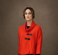 A woman with shoulder-length hair stands against a plain background, wearing a bright red coat with front buckles and a high collar. Beneath the coat, her timeless look is complemented by layering the cozy Long Sleeve Knit 6374, a 100% cotton sweater from MARBLE.