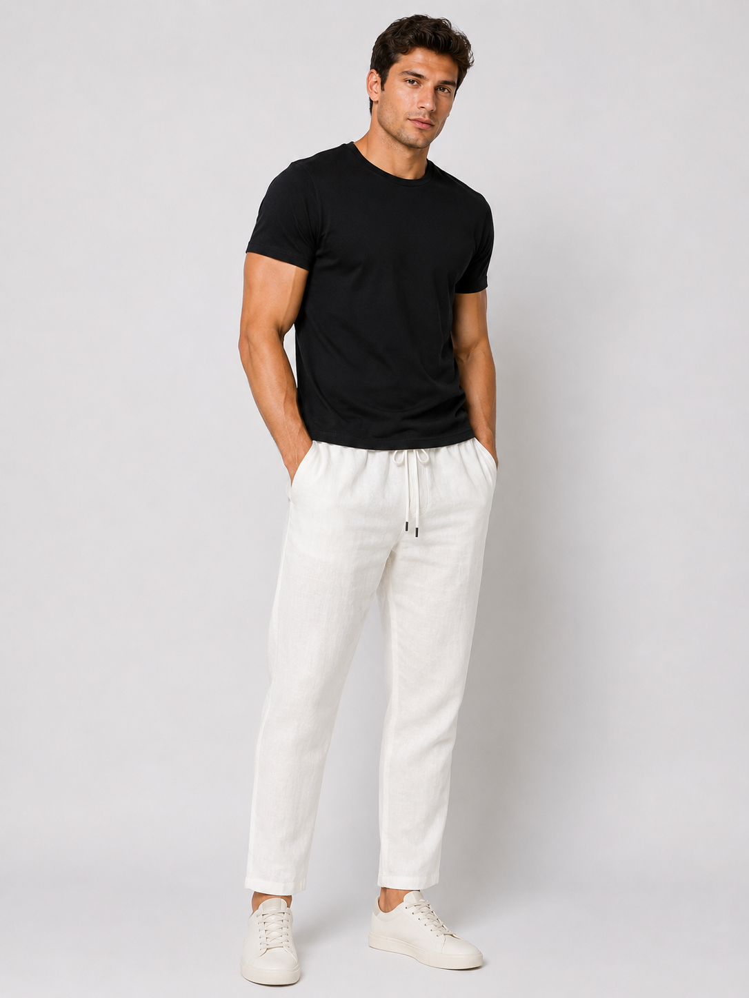 A man stands against a plain light gray background wearing a black short-sleeve t-shirt, white sneakers, and Paul Miranda’s Linen Pant White PE618JK with a white drawstring, hands in his pockets and displaying a neutral expression.