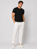 A man stands against a plain light gray background wearing a black short-sleeve t-shirt, white sneakers, and Paul Miranda’s Linen Pant White PE618JK with a white drawstring, hands in his pockets and displaying a neutral expression.