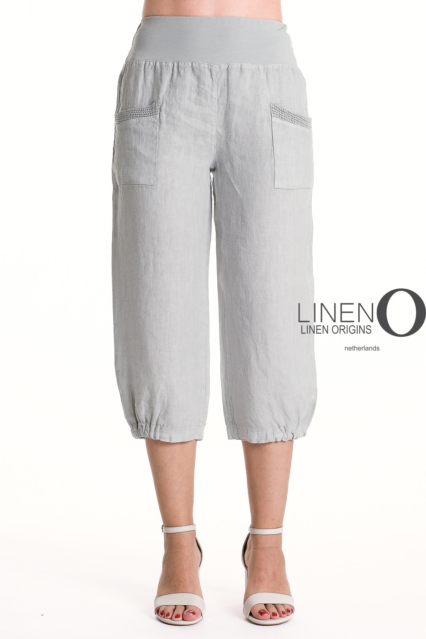 A person wears LinenO's French Linen Capri PT878-152 in Grigio Chiaro with elastic waist and cuffs, front pockets, and pairs them with light gray heeled sandals. The "Linen Origins Netherlands" logo is visible.