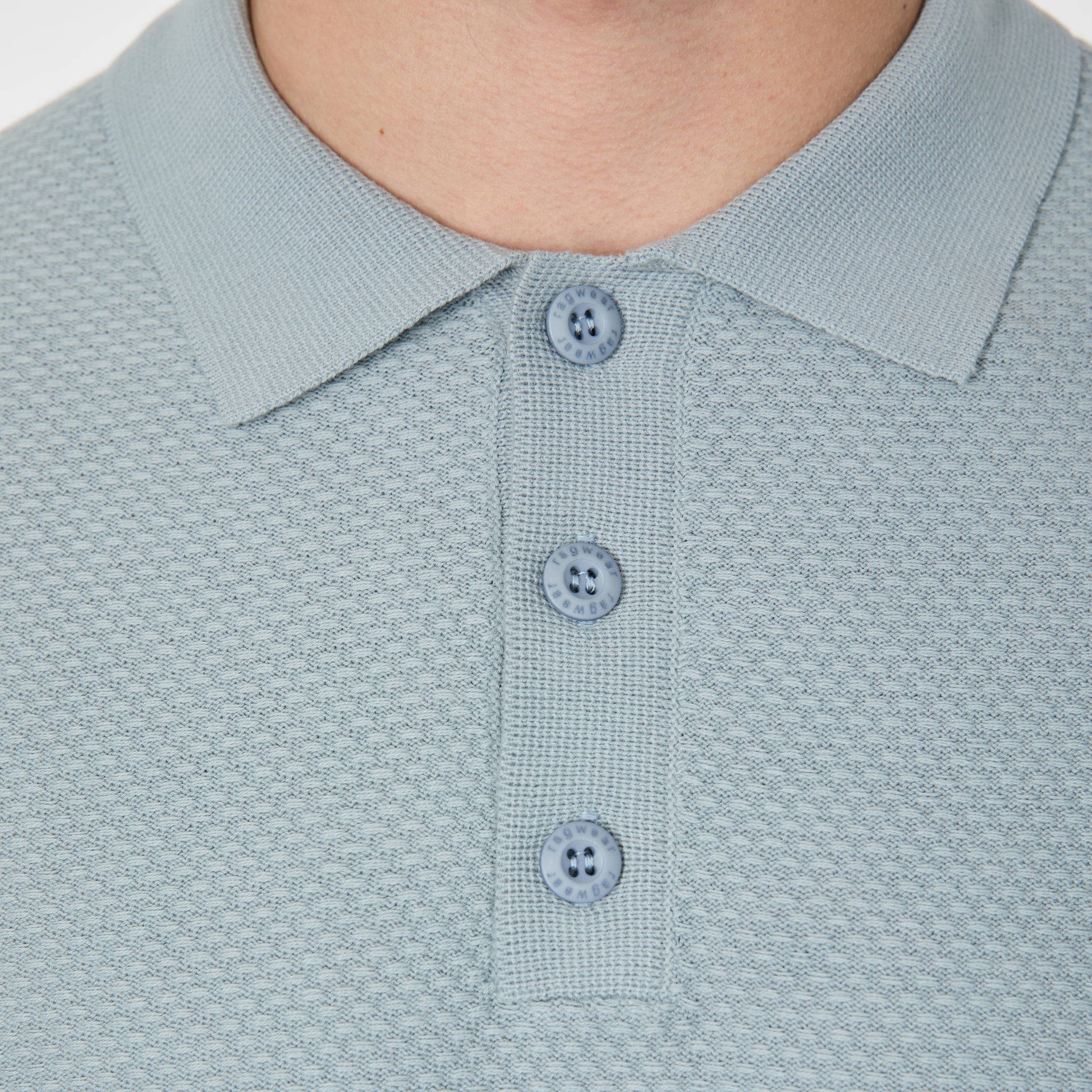 Close-up of a person wearing the RAGWEAR Polo Tarsh Short 2612-15050-2046 in Dusty Blue, featuring textured light blue 100% cotton fabric, three matching buttons, and a classic collar. Only the neck and upper chest are shown.