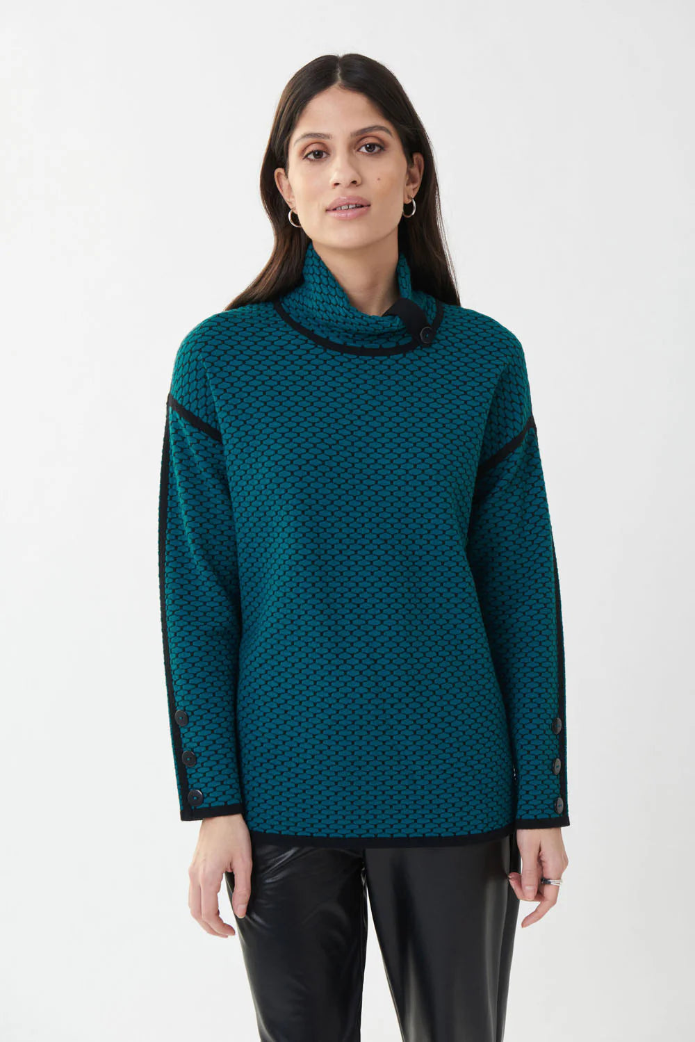 A woman with long dark hair is wearing a Joseph Ribkoff Blue Lagoon Textured Knit Top (Style 223953) in teal-blue, accented with black shoulders and buttons on the sleeves. She pairs it with black pants and stands against a plain white background.