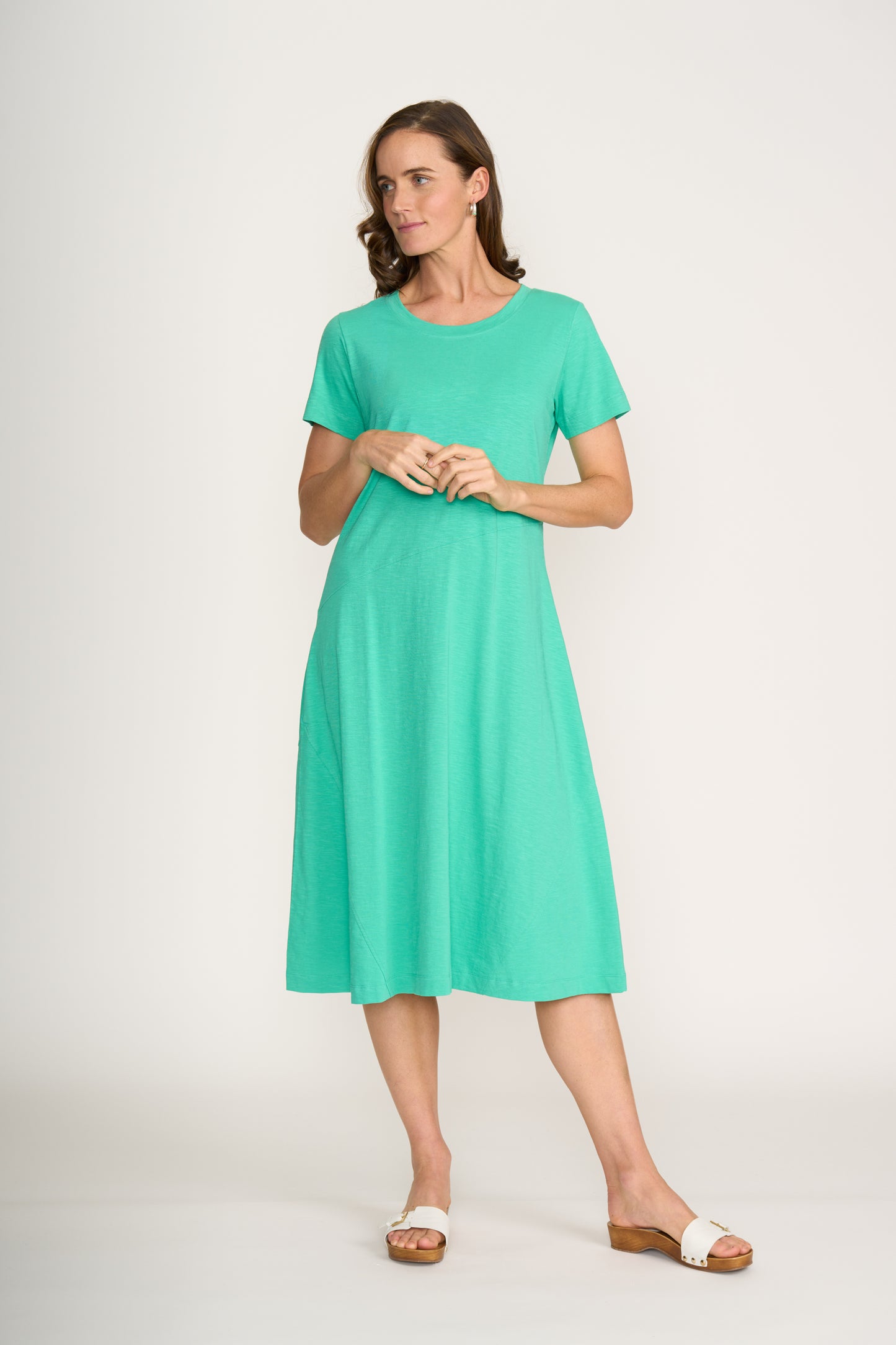 A woman wearing the FOIL Stitch Together Dress FO8181 in Aquamarine stands against a plain white background, looking to the side with her hands clasped in front of her. She pairs the dress with white sandals.