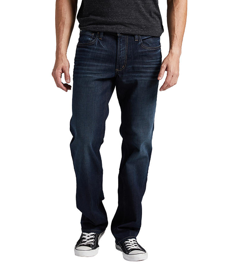 A person wearing Allan Slim Fit Straight Leg Jeans by Silver Jeans Co., a dark shirt, and black-and-white sneakers stands against a plain white background. The person's arms hang naturally by their sides, with the left arm slightly bent. Only the lower half of the body is visible.