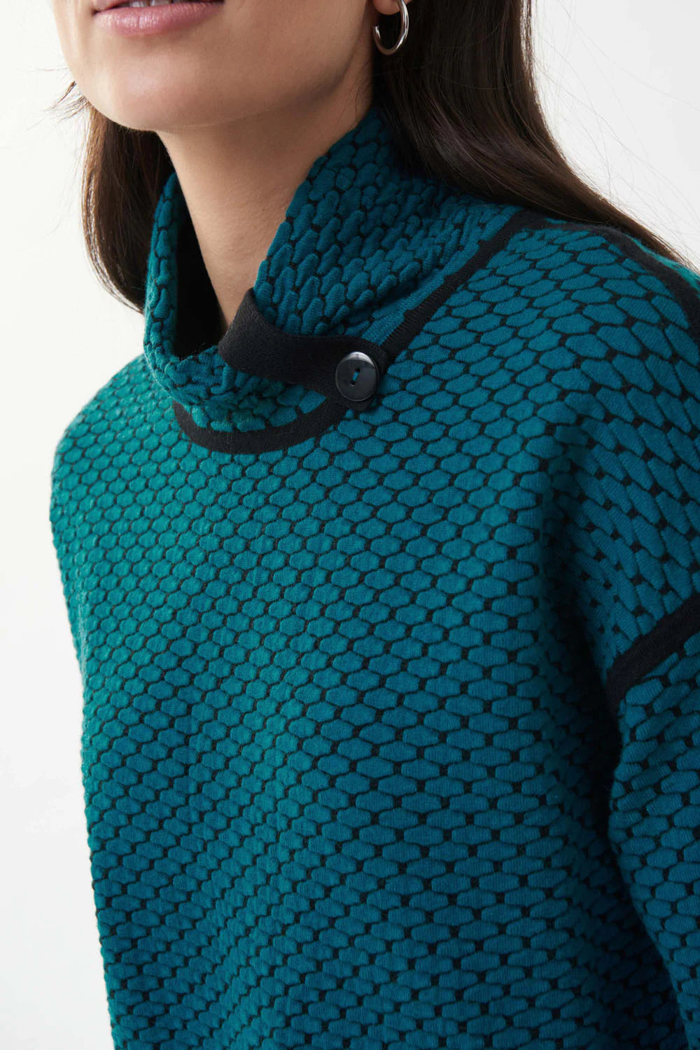 A woman with long dark hair is wearing a Joseph Ribkoff Blue Lagoon Textured Knit Top (Style 223953) in teal-blue, accented with black shoulders and buttons on the sleeves. She pairs it with black pants and stands against a plain white background.