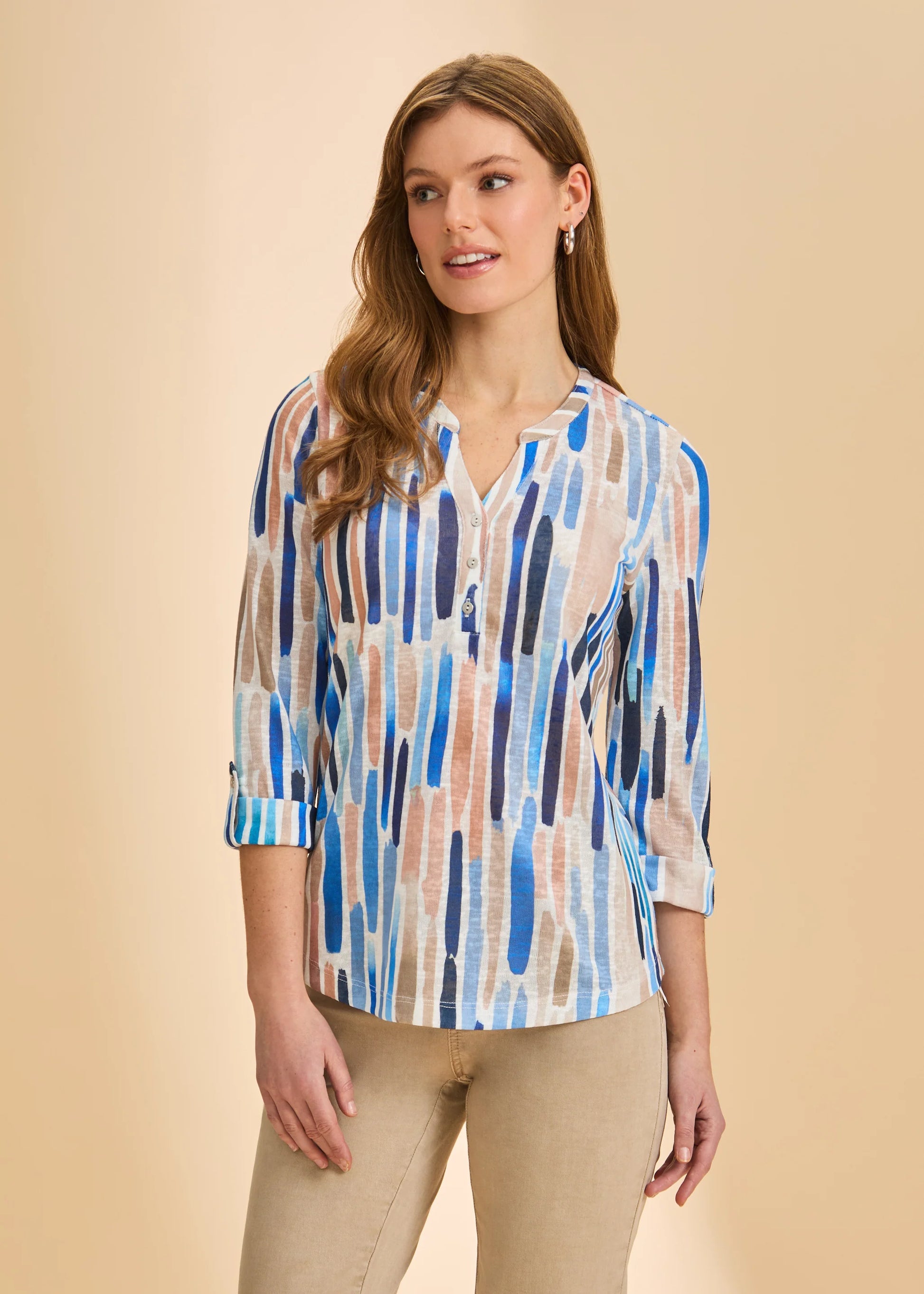 A woman wears the French Dressing Jeans 3/4 Sleeve Henley Top in Zippy Neutral, featuring a blue, beige, and brown brushstroke pattern with beige pants, standing against a plain beige background and smiling softly.