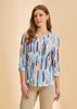 A woman wears the French Dressing Jeans 3/4 Sleeve Henley Top in Zippy Neutral, featuring a blue, beige, and brown brushstroke pattern with beige pants, standing against a plain beige background and smiling softly.