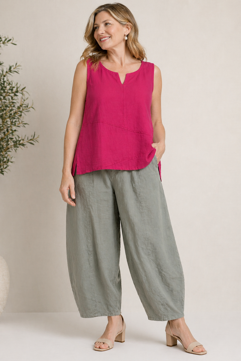 A woman smiles in the TaraVao Sleeveless Top Marina 2094, a bright pink notched-neck top, paired with loose light gray linen-cotton wide-leg pants, sandals, and loose wavy hair.