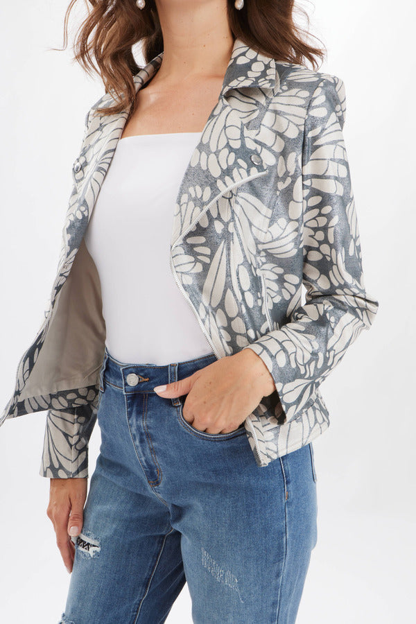 A woman with long brown hair is wearing a Joseph Ribkoff Printed Moto Jacket Style 231911, which features a butterfly-inspired motif, over a white top and blue jeans. She is smiling and looking at the camera while holding the jacket's lapel. The background is plain white.