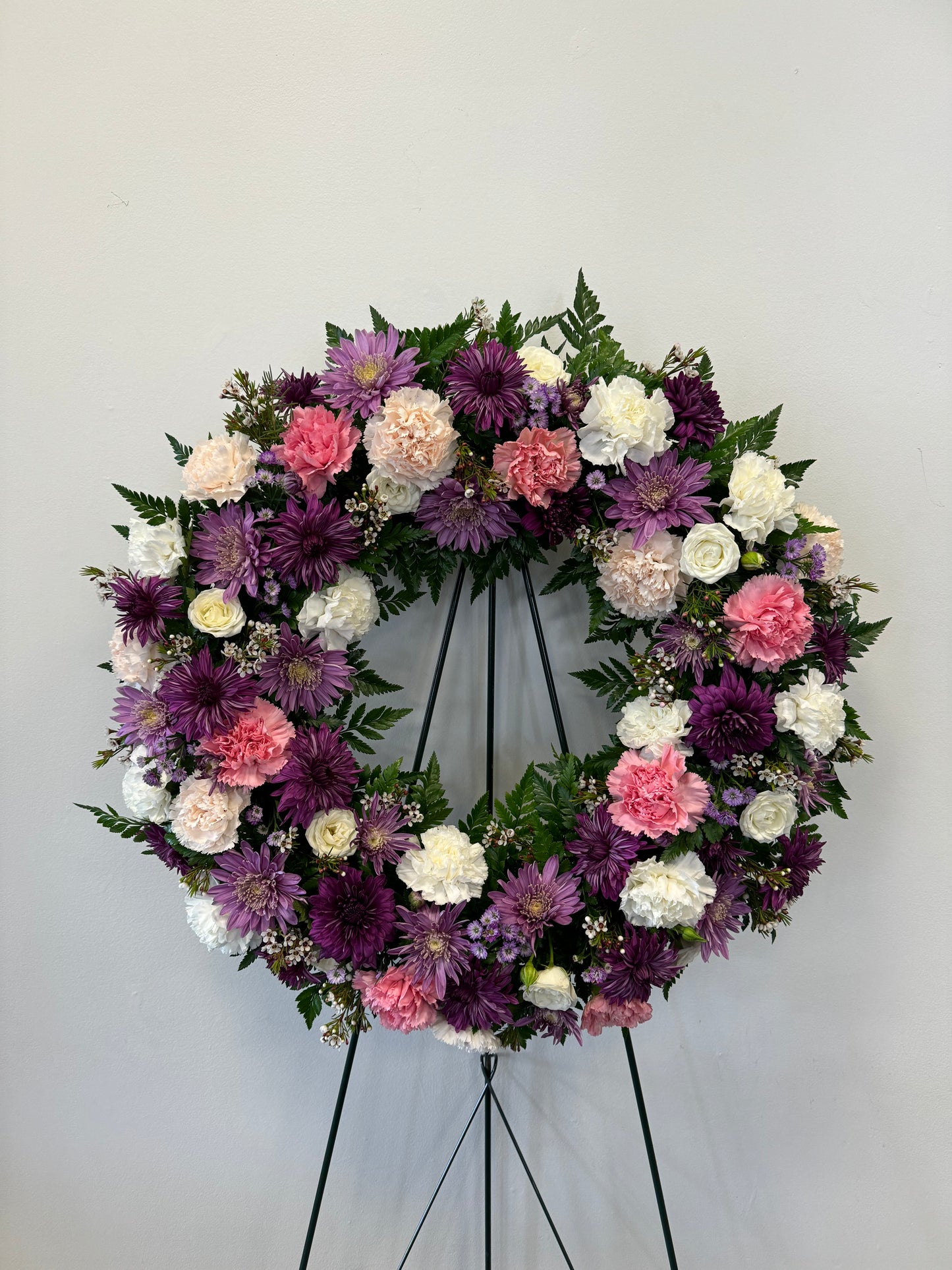 The "Soft Embrace Wreath" by Posies Flowers & Fashion is a large, circular wreath on a stand, beautifully adorned with a vibrant mix of purple, pink, and white flowers in full bloom, and accented with lush green foliage. It is displayed against a plain, light-colored wall. Please note that availability may vary based on seasonality.