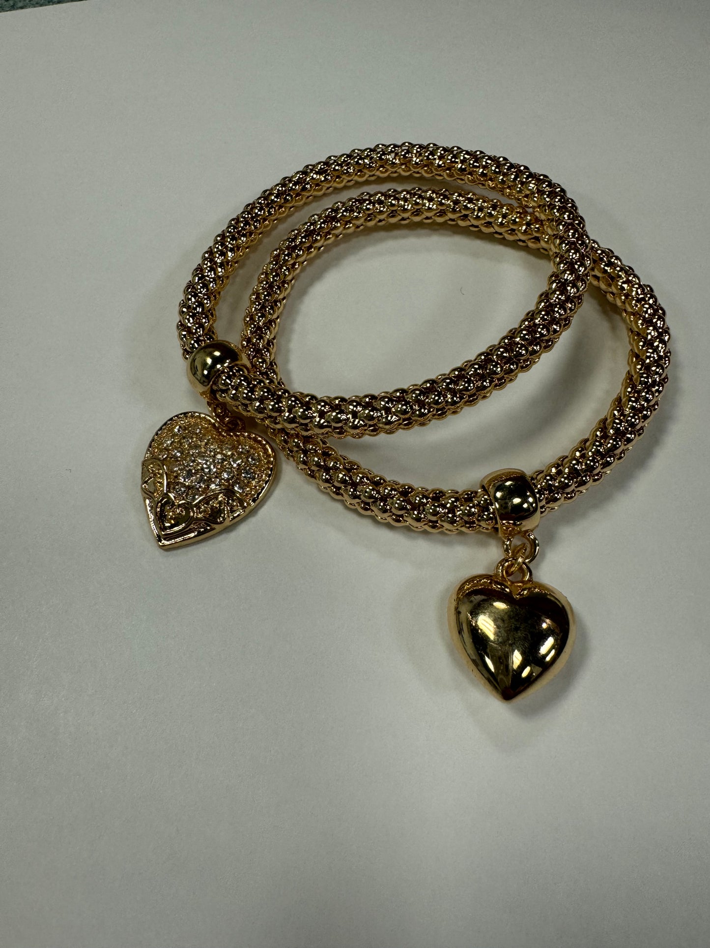 Two Gina 33 Bracelets from Elise Accessories, each featuring gold braided designs and heart-shaped pendants, rest elegantly on a plain white surface. One pendant boasts a textured finish while the other is polished and shiny—a perfect duo for any heart charm bracelet enthusiast.