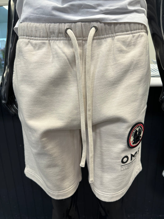 A mannequin is wearing the White Sand Yabu Shorts from Once We Were Warriors, featuring an embroidered patch and a logo on the left thigh. These fleece shorts have an elastic waistband with a visible drawstring. The mannequin is set against a dark background.