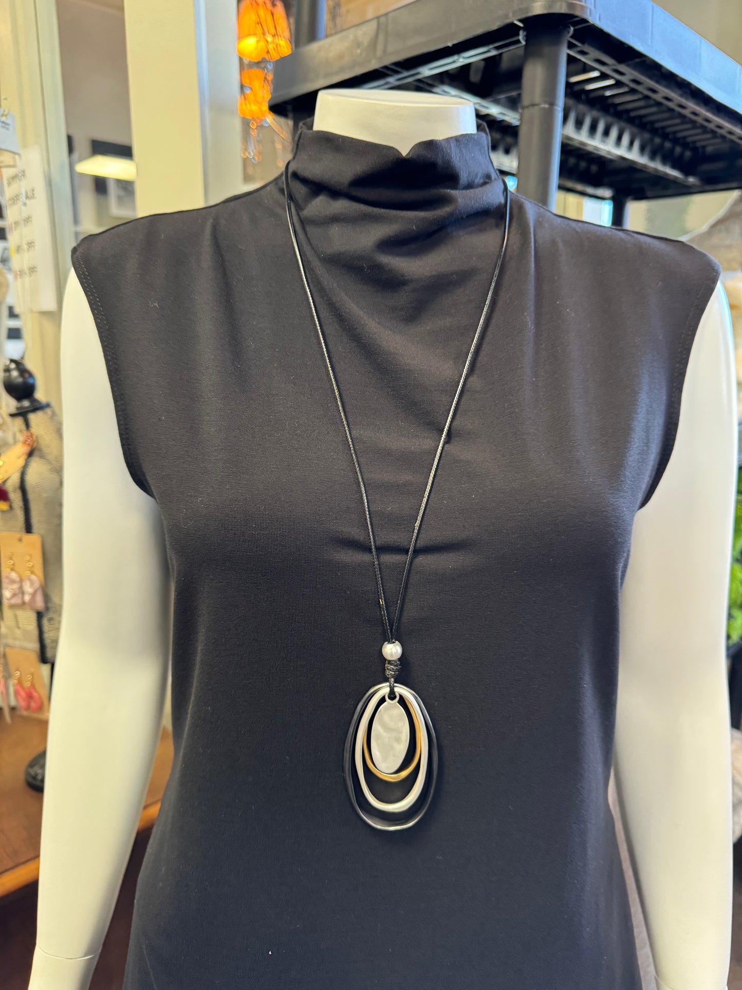 A mannequin dressed in a sleek black sleeveless turtleneck dress is adorned with the stunning Sofia 10 Necklace from Elise Accessories, featuring three oval pendants in black, silver, and white. The blurred indoor setting provides an elegant backdrop to this striking jewelry piece.