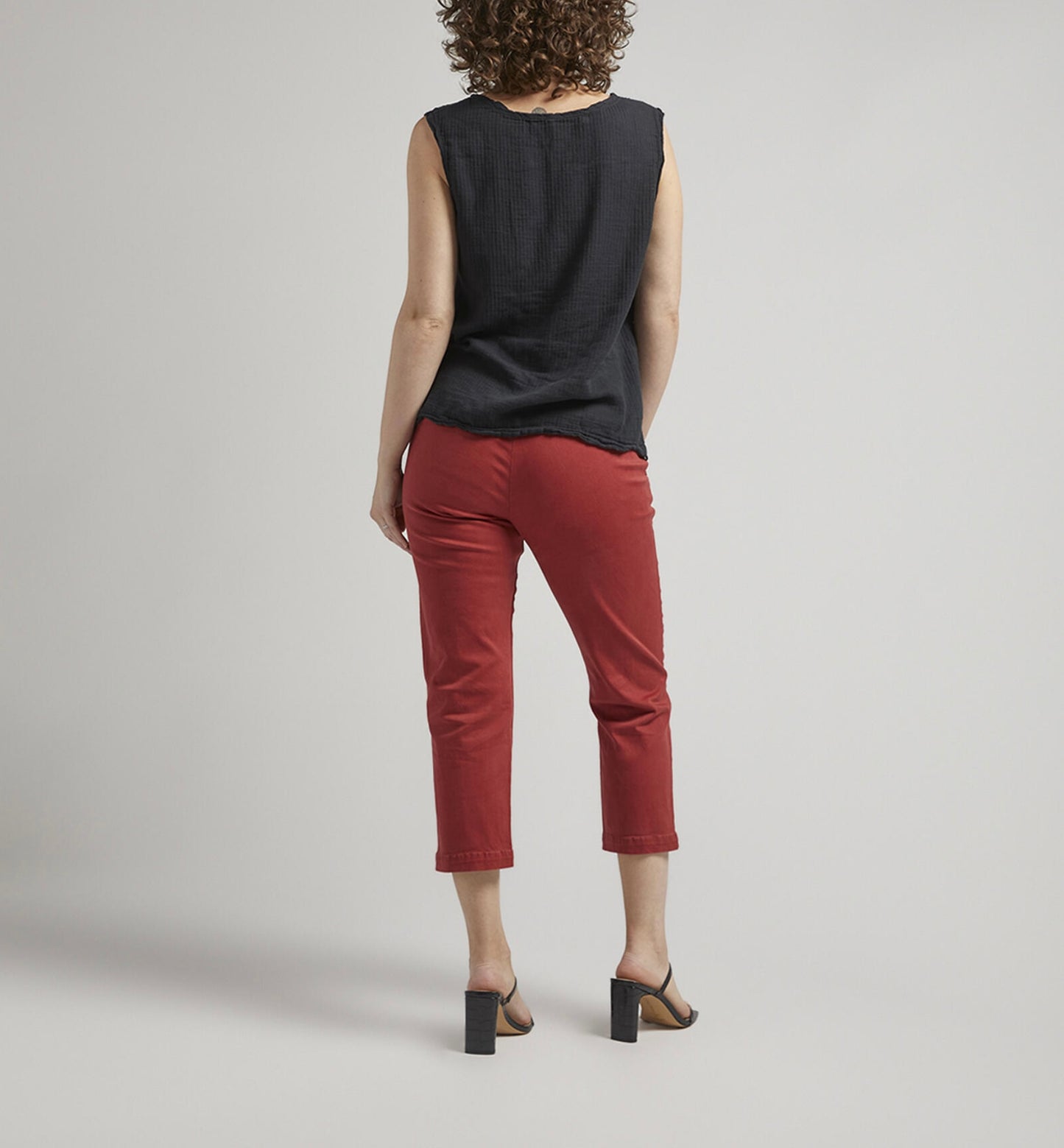 A person stands against a plain backdrop with a neutral expression. They are wearing a sleeveless black top, flattering Maddie Capri Mid Rise pants in brick-red from JAG, and black heeled sandals. Their left hand rests in their pocket, and they have curly hair.