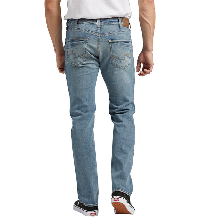 A person is standing sideways, dressed in Allan Slim Fit Straight Leg Jeans from Silver Jeans Co., a white T-shirt, and black slip-on shoes. The jeans have a fitted silhouette with slight fading on the thighs and knees. The person's arm is relaxed by their side.