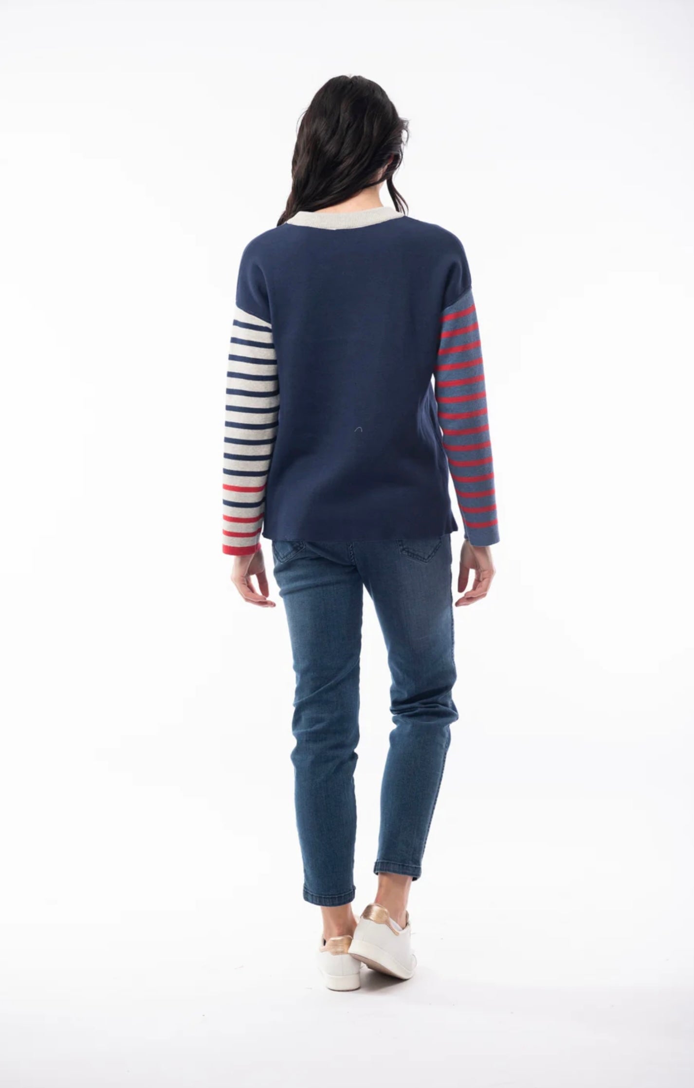 A woman with long dark hair is wearing the ORIENTIQUE - REVERSIBLE ROUND NECK SPOT/STRIPE JUMPER (1229) made of organic cotton, featuring a navy blue background adorned with white polka dots and red striped sleeves. She's paired it with blue jeans and white sneakers, and stands in a casual pose, slightly leaning back with her hands behind her. The background is plain white.