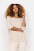 A woman with curly hair models the versatile Knitted Top 26244-40 by SoyaConcept, a lightweight beige sweater styled with white pants. She poses confidently, one hand on her hip and the other touching her hair against a plain white background.
