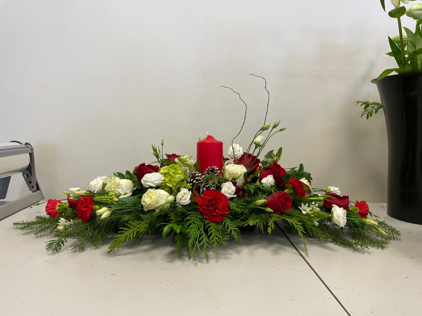 The Pillar Candle Centrepiece by Posies Flowers & Fashion features red and white roses, lush greenery, and a striking red pillar candle in the center, beautifully arranged on a white surface against a plain background.