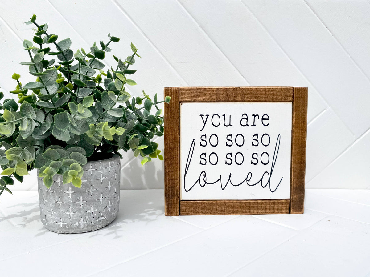 A potted plant sits next to the Inspired Findings “You Are So So Loved Mini” sign, which features an antique walnut frame and displays “you are so so so so so so loved” in black cursive and print letters on a white background.