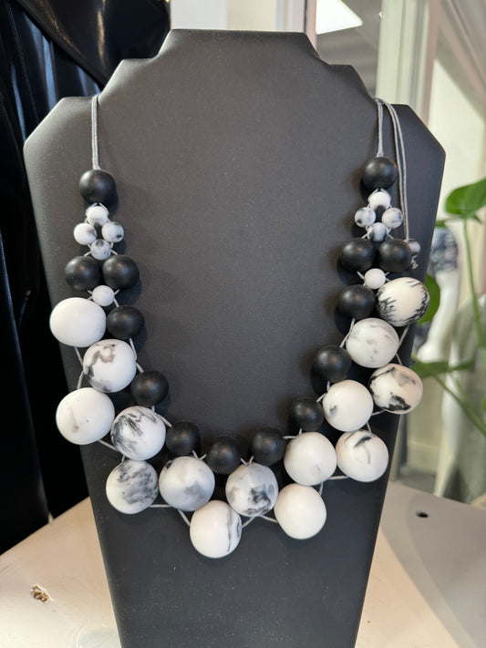 A Suzie Blue Black and White Beaded Necklace displayed on a black bust. This elegant accessory consists of graduated large beads, blending solid black with marbled white. In the background, there's a glimpse of a potted plant and parts of a store interior.