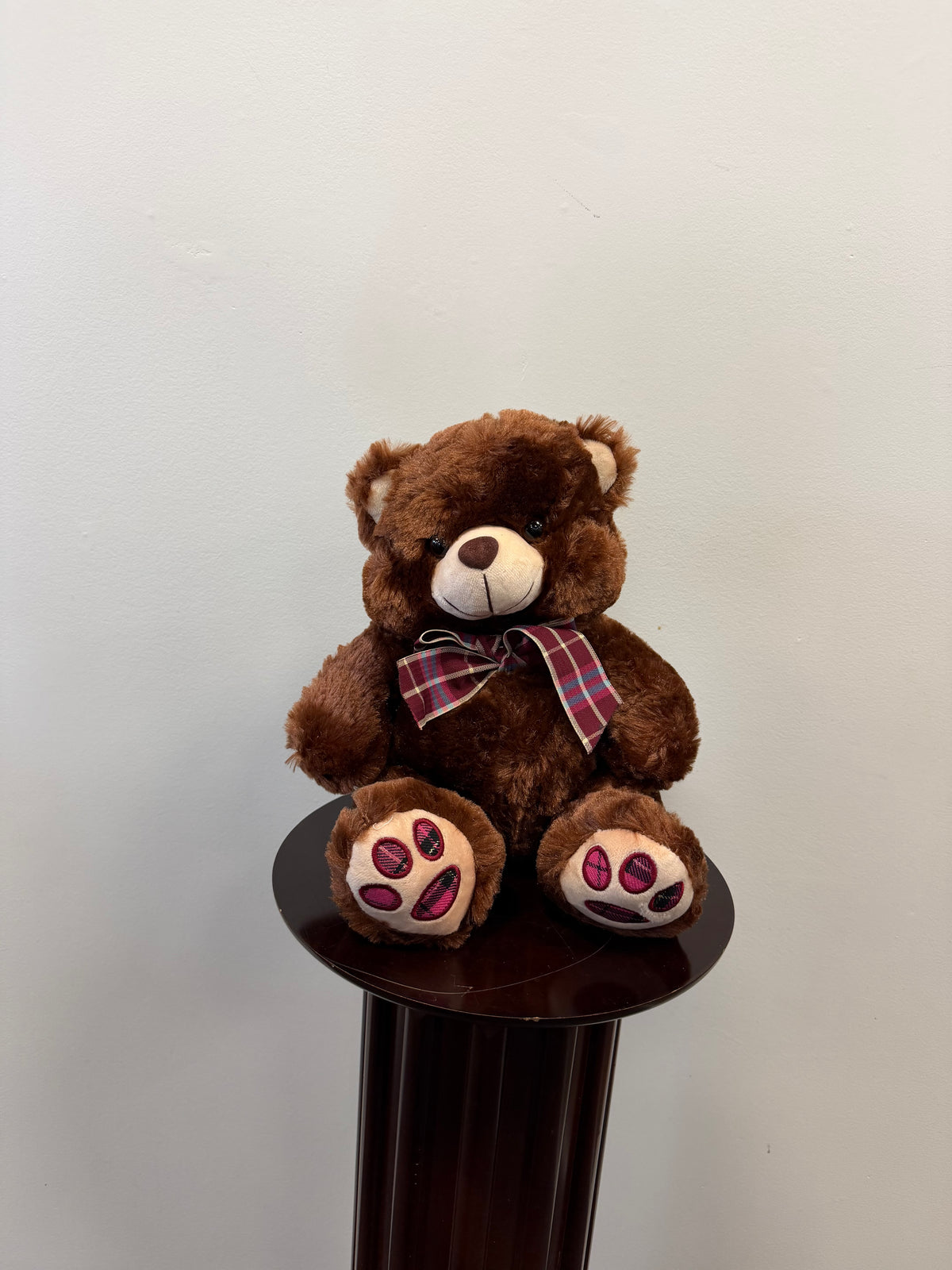 A Sproule Brown Teddy Bear with a plaid bow sits on a dark wooden pedestal against a plain light-colored wall, its pink paw pads adding a charming touch.