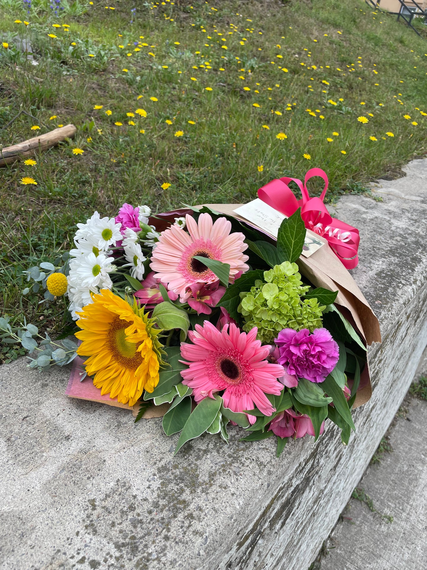 A stunning bouquet called Designer’s Choice Freshcuts from Posies Flowers & Fashion is resting on a white surface. The arrangement features vibrant orange and pink roses, cheerful yellow chrysanthemums, elegant white flowers, and assorted green foliage. It is partially wrapped in brown paper and elegantly tied with a burgundy ribbon.