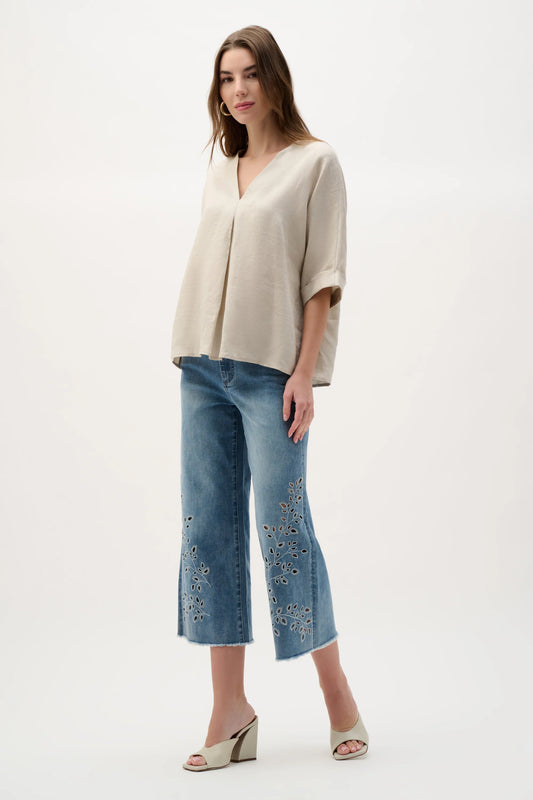 A woman stands against a plain background wearing the Joseph Ribkoff Linen Straight Top With V-notch neckline in moonstone, paired with blue embroidered cropped jeans and cream open-toe wedge sandals.