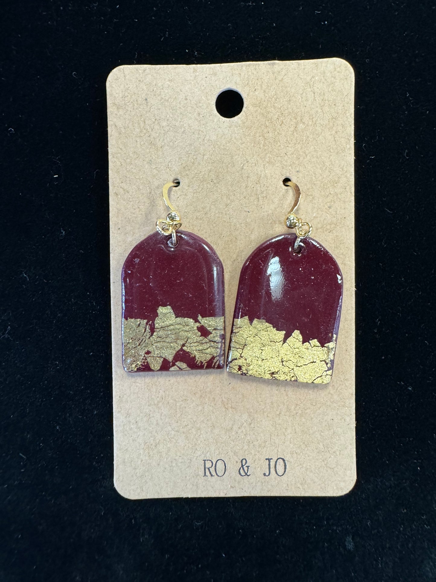 A pair of Ro&Jo Earrings 424 from the RO&JO brand are displayed on a brown cardboard backing labeled "RO & JO." These handmade earrings feature red polymer clay with gold foil flakes embedded in the bottom half, and they are attached to gold hooks. The background is black.