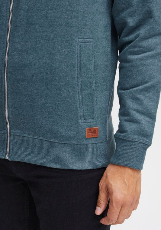 A person wearing the Ragwear Sweatshirt Zip - TRAYNE TURQUOISE by RAGWEAR is shown from the shoulders to the mid-thigh. Their right hand is visible, resting by their side. The turquoise sweatshirt features a small brown patch with writing near the bottom hem, just above their dark-colored pants.