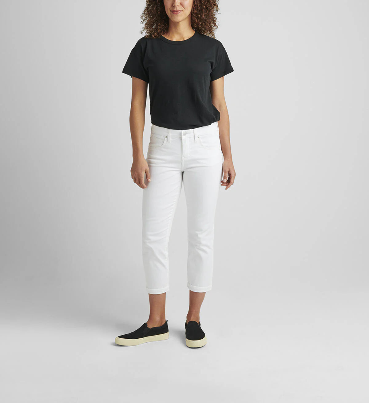 A person with curly hair, wearing a black t-shirt and the Cecilia Mid Rise Capri White from JAG, paired with black sneakers with white soles, is standing and facing away from the camera. The background is plain and gray.