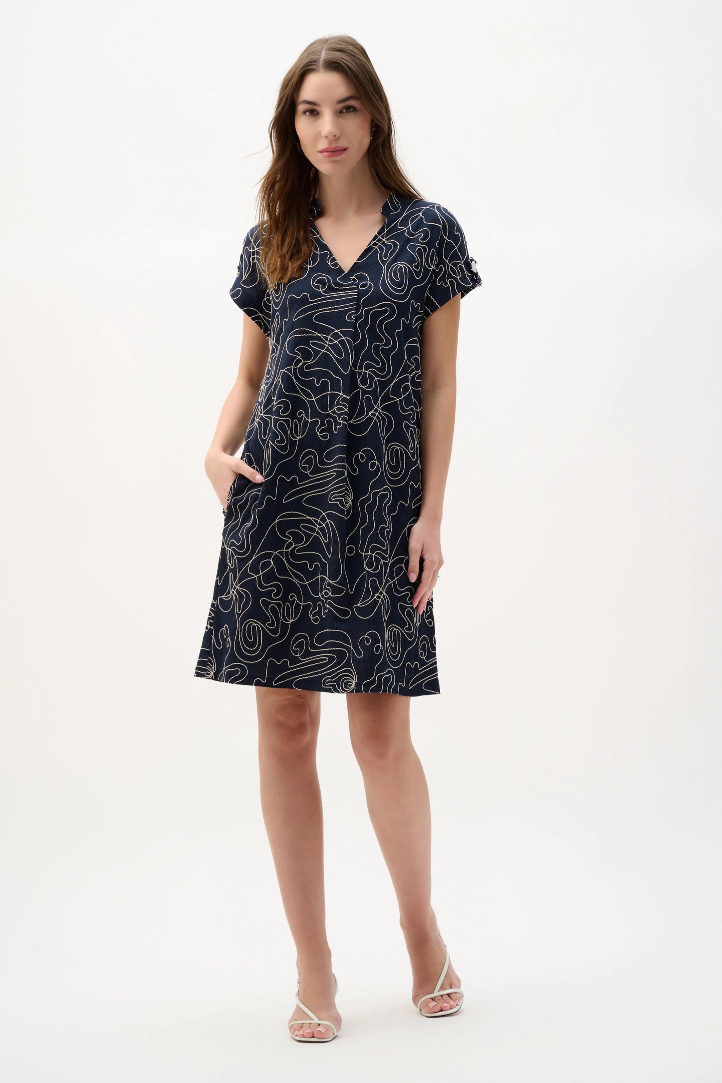 A woman models the Joseph Ribkoff Novelty Abstract Print Trapeze Dress 262170, featuring short dolman sleeves and a V-neckline pleat detail, with one hand in her pocket and the other by her side against a plain white background.