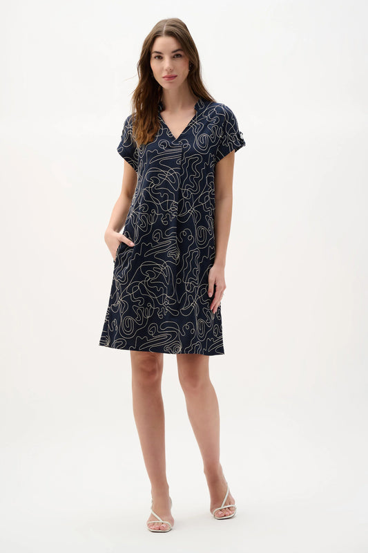 A woman models the Joseph Ribkoff Novelty Abstract Print Trapeze Dress 262170, featuring short dolman sleeves and a V-neckline pleat detail, with one hand in her pocket and the other by her side against a plain white background.