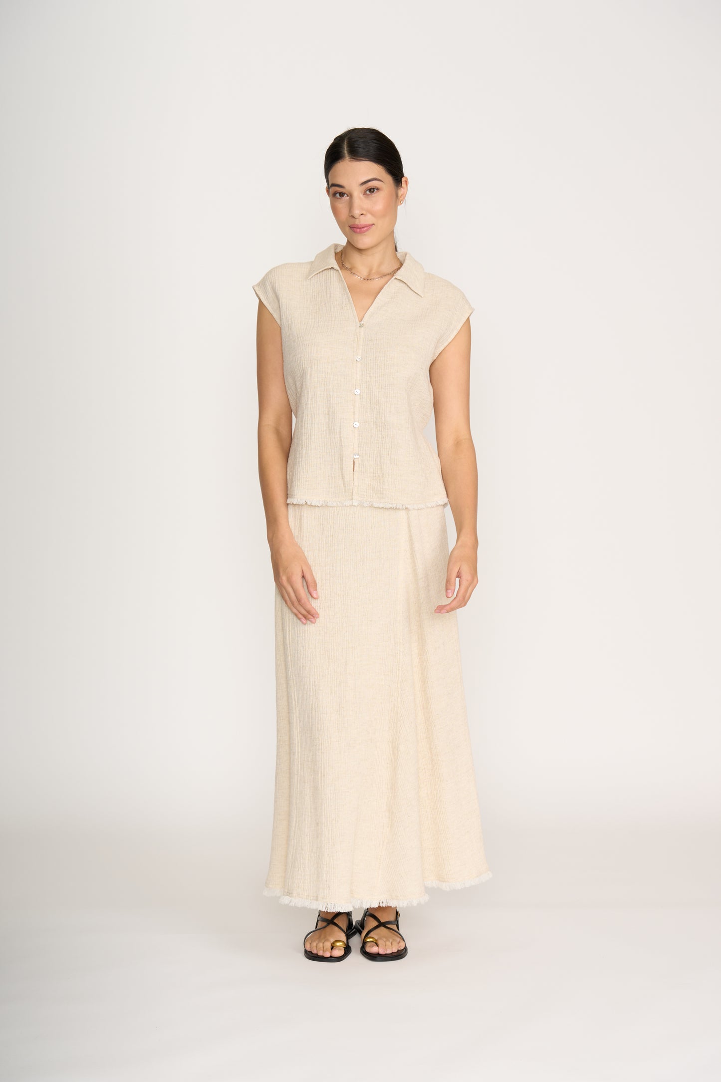 A woman with dark hair pulled back stands against a white background, wearing the FOIL Natural Blender Shell Sandshell FO8474—a lightweight linen-rayon blend button-up top with a matching long skirt featuring frayed hems—and black sandals.