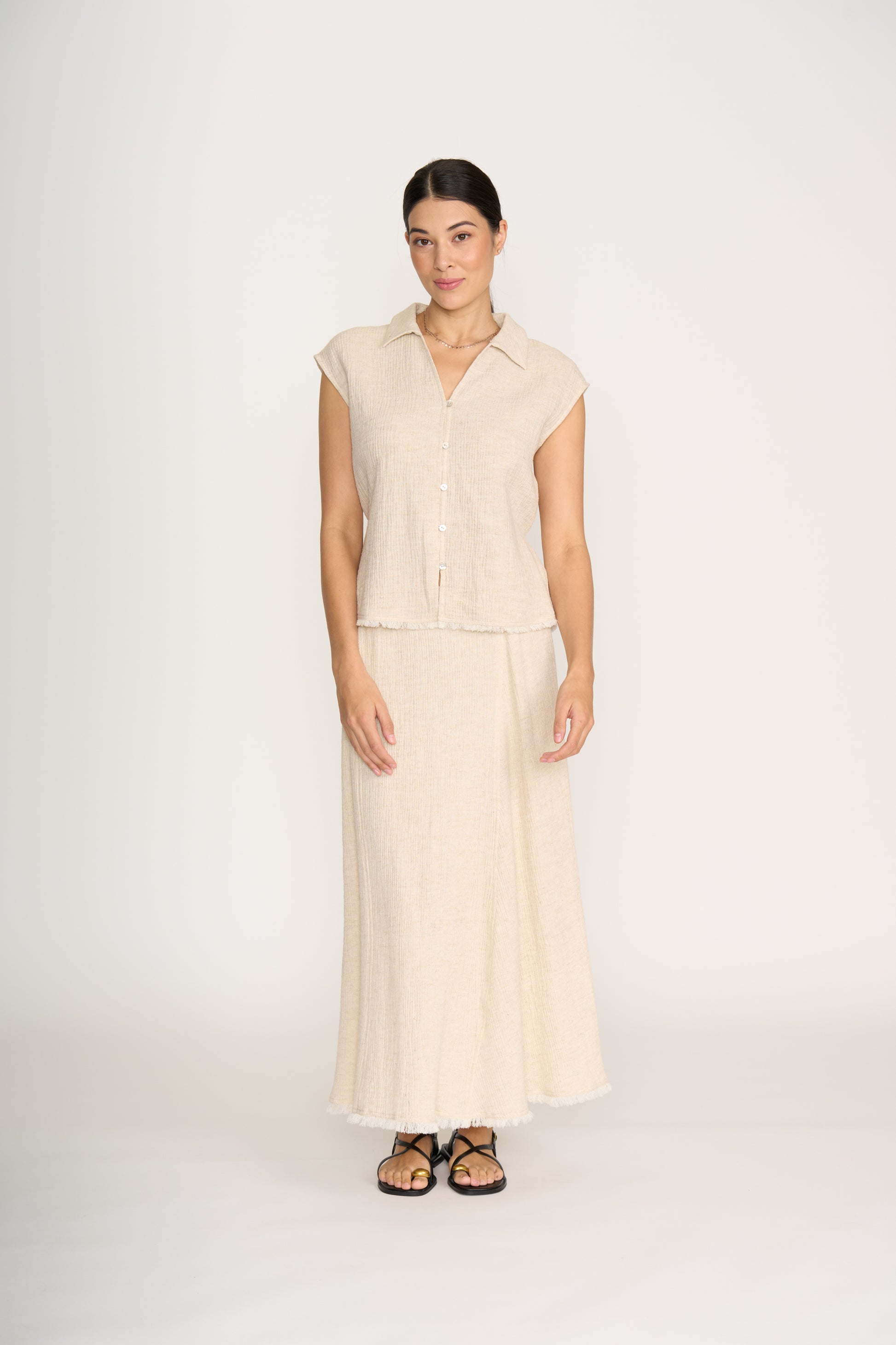 A woman with dark hair pulled back stands against a white background, wearing the FOIL Natural Blender Shell Sandshell FO8474—a lightweight linen-rayon blend button-up top with a matching long skirt featuring frayed hems—and black sandals.