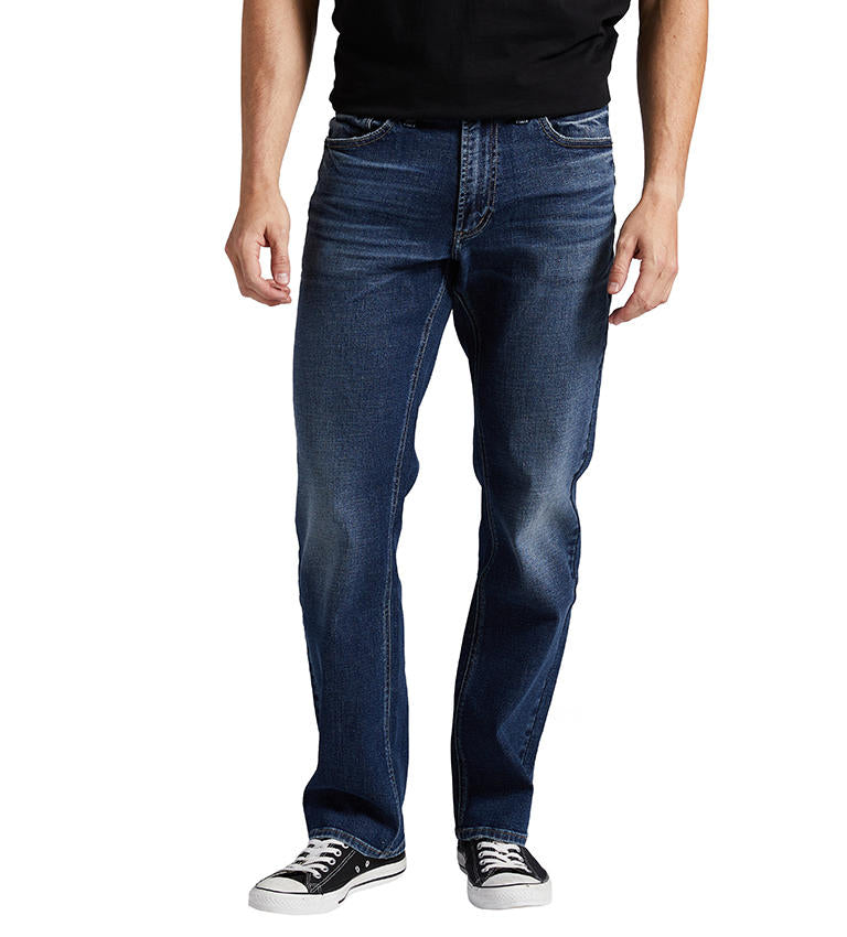 A person is standing, wearing Silver Jeans Co.'s Grayson Classic Fit Straight Leg Jeans in a dark indigo wash with classic five-pocket styling. The slightly faded straight-leg jeans are paired with a black shirt and black low-top sneakers with white soles. The background is white.