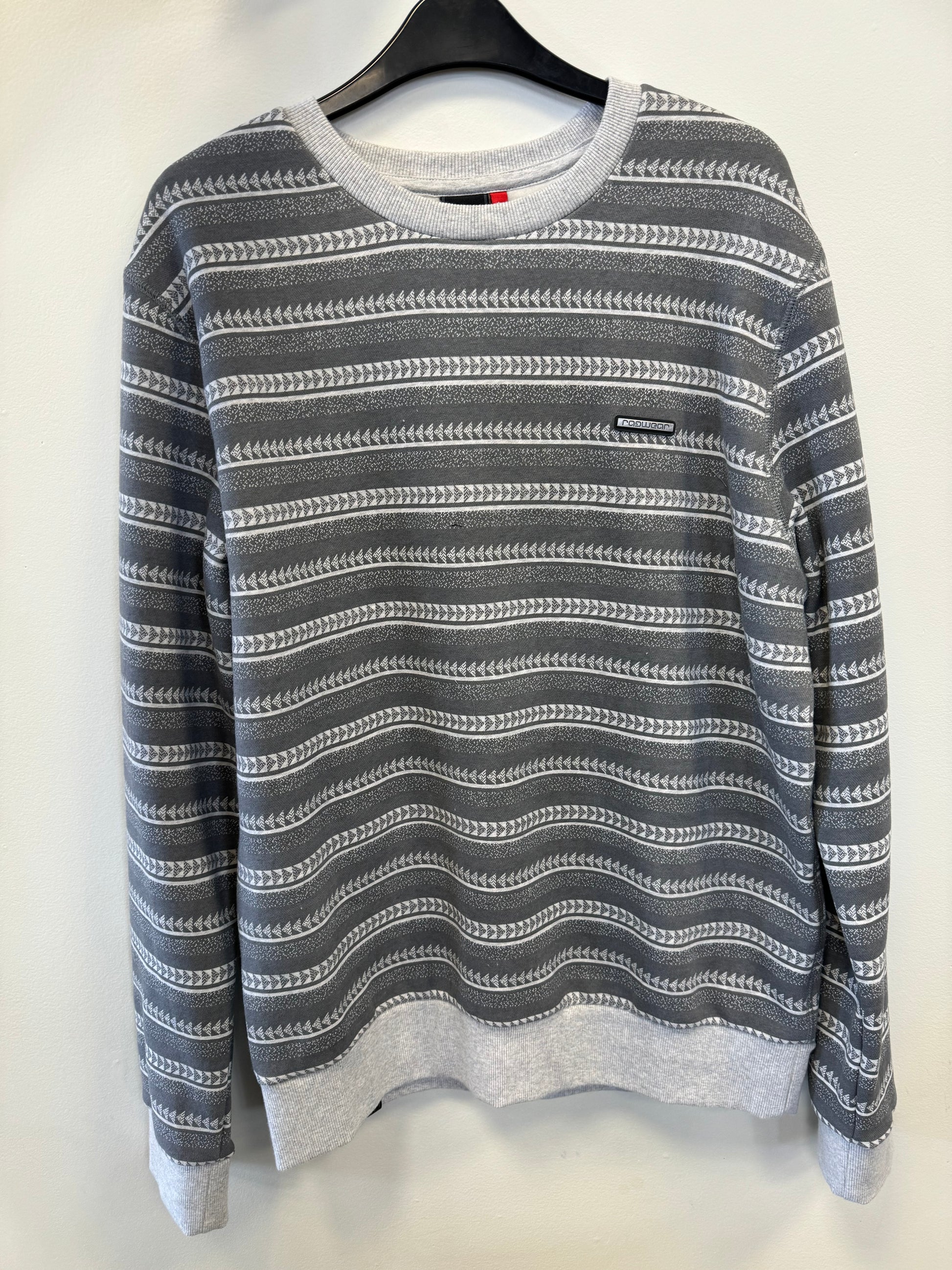 A Ragwear Sweatshirt Pullover - INDDIE PRINT by RAGWEAR, featuring gray and white stripes with a pattern of horizontal wavy lines and white geometric shapes, is hanging on a black hanger against a white wall. The vegan sweatshirt also has gray cuffs and hem, and a small black tag with writing is sewn on the front.